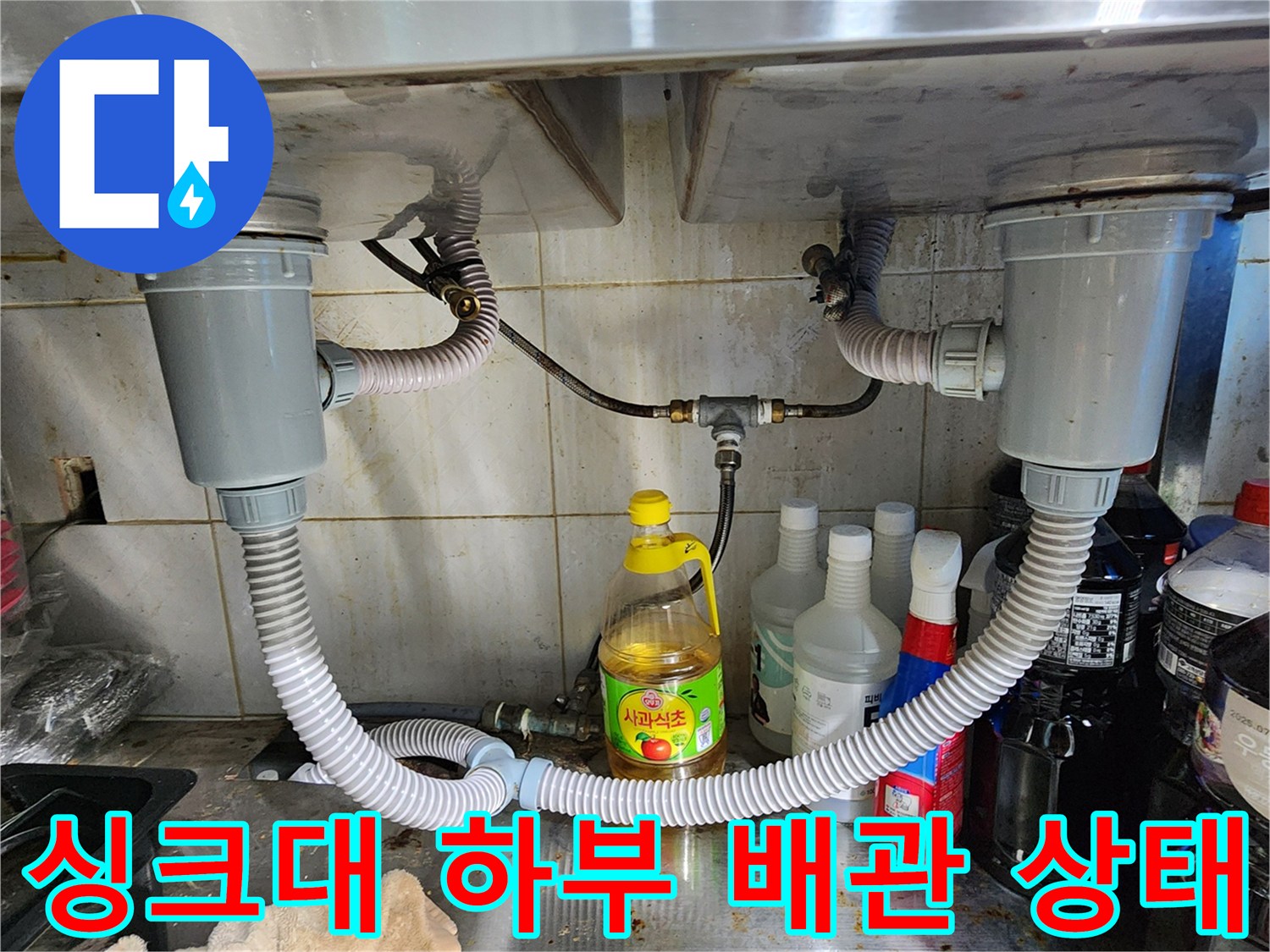 싱크대 아래쪽의 굵은 배수 호스와 금속 재질의 배관 연결 장치입니다.