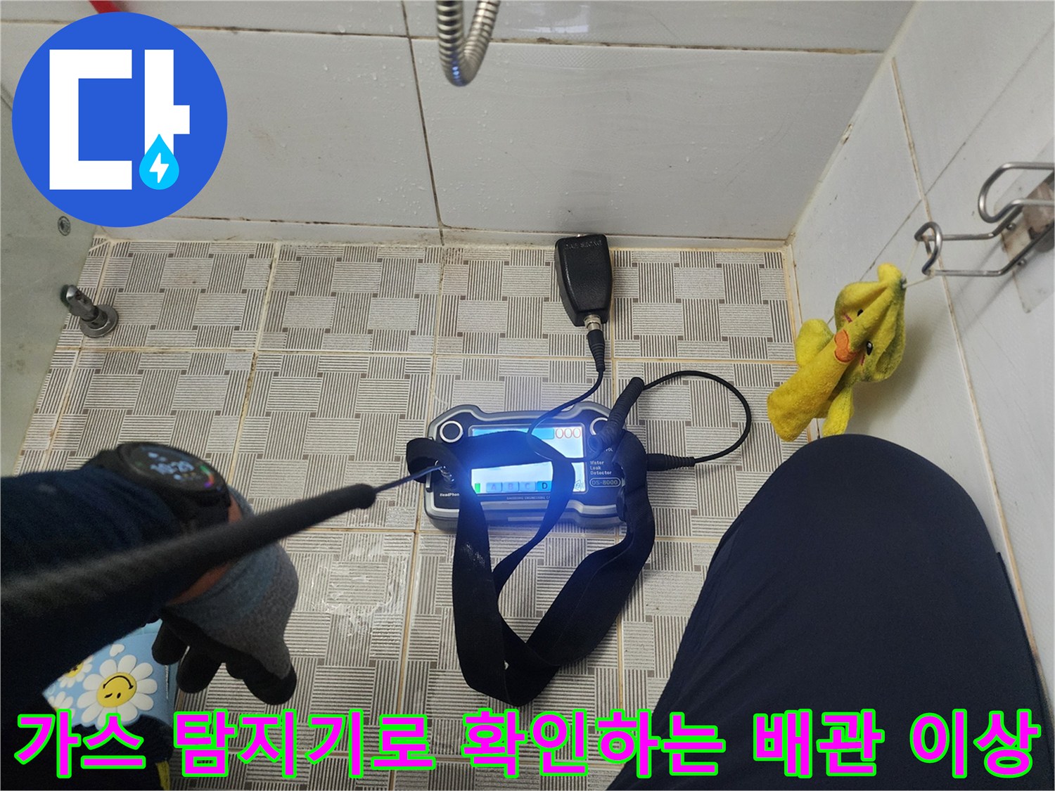 욕실 바닥 타일 위에서 가스 탐지 장비를 사용하여 누수 지점을 찾는 장면입니다.