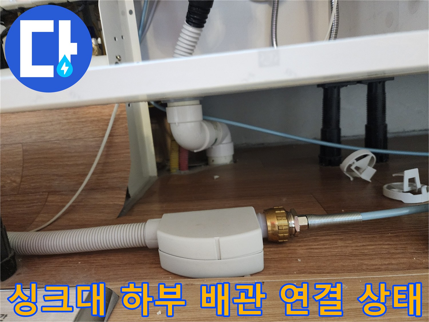 주방 싱크대 아래쪽의 굵은 주름관과 금속 재질의 배관 연결 부위 모습입니다.