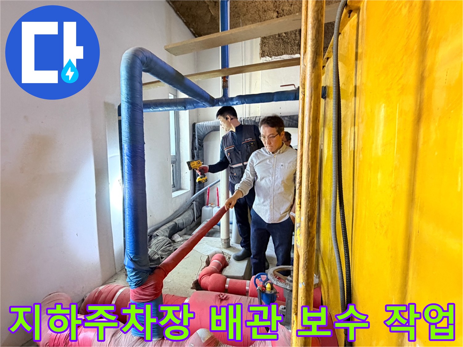 지하 주차장 천장 내부의 파란색 및 분홍색 보온재로 감싸진 배관을 엔지니어가 점검하고 있습니다.