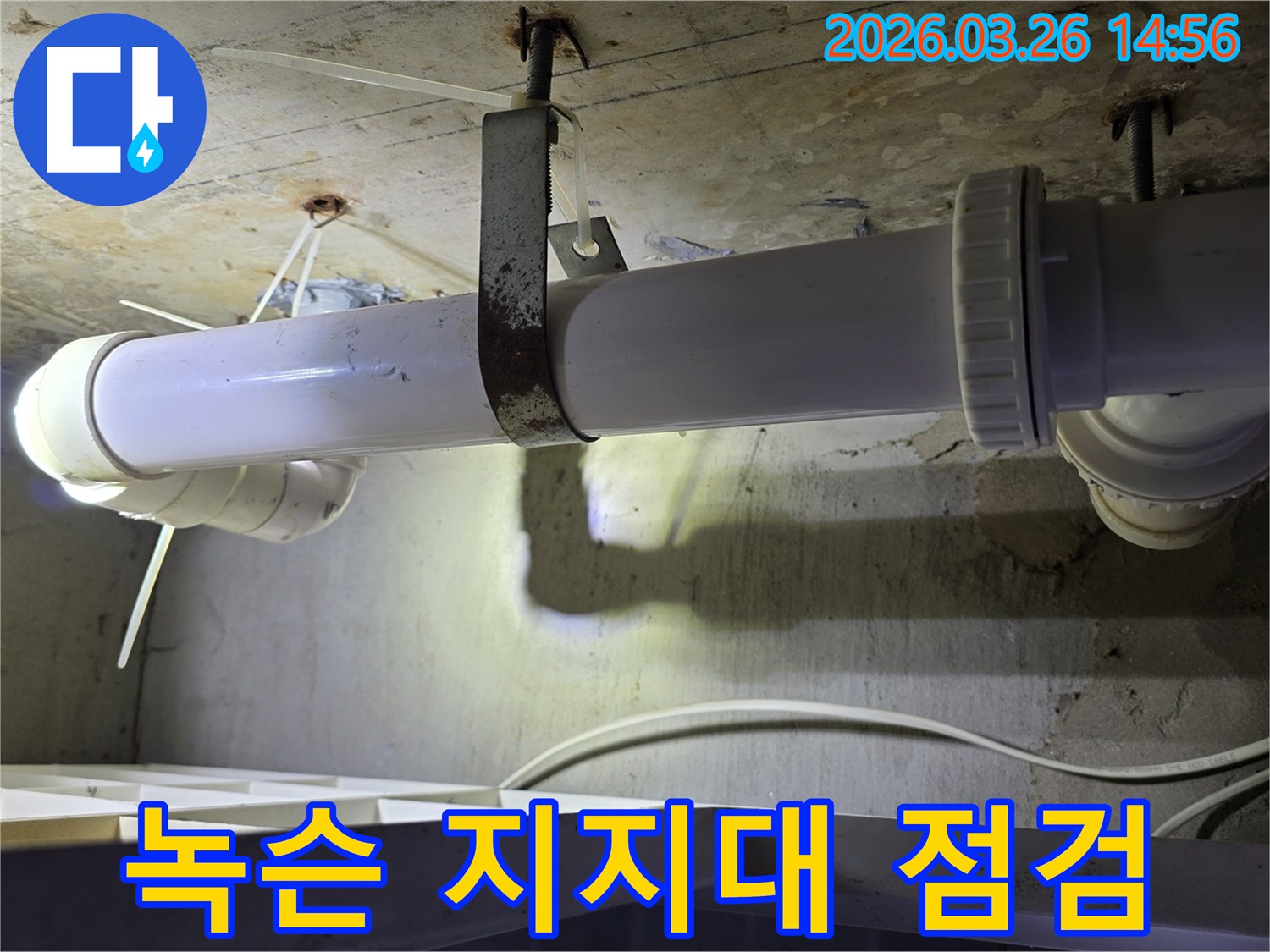 콘크리트 천장에 고정된-녹이 심하게 슨 금속 배관 지지대와-그 아래로 지나가는 하얀색 배관의 모습