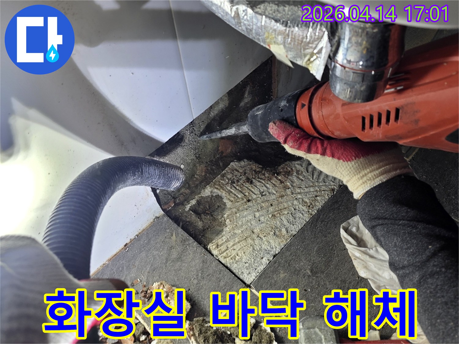 누수 지점의 바닥 타일을 해체하는 작업자가 드릴을 사용하며-파손된 타일 조각과 먼지 흡입 호스가 함께 보이는 작업 현장