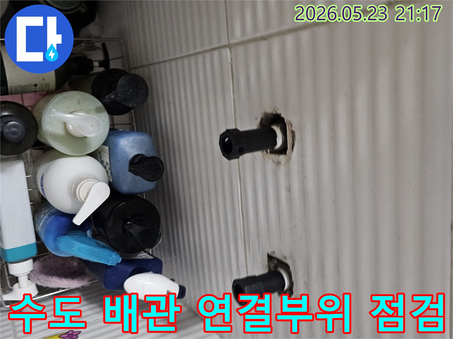 타일 벽면에서 돌출된 두 개의 수도 배관 연결 부위를-누수 발생 여부를 확인하기 위해-면밀히 검사하는 모습입니다.