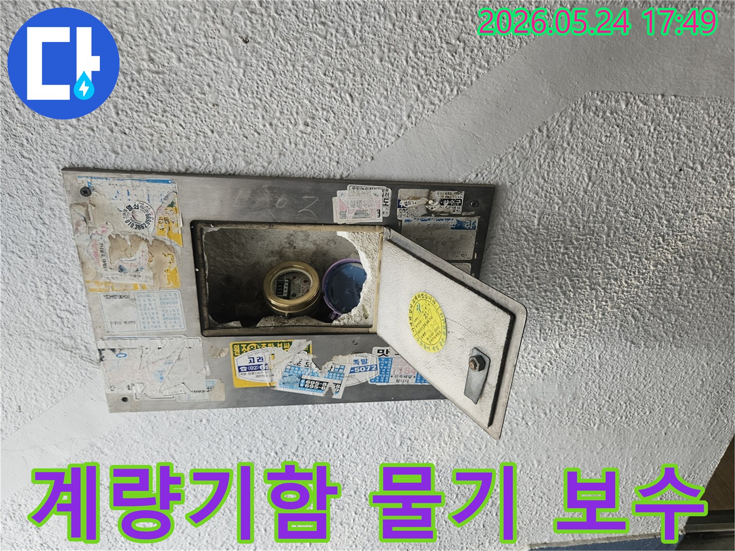 빌라 외벽의 수도계량기함이 열려있고-내부에 물기가 확인되어-보수 작업이 필요한 상태를 보여줍니다.