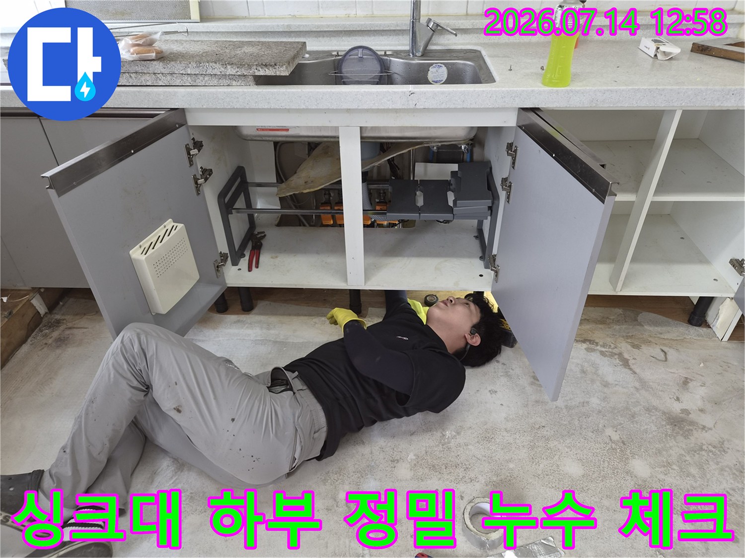 작업자가 바닥에 누워 싱크대 배수 시스템의 연결 상태를 점검하고 있는 장면