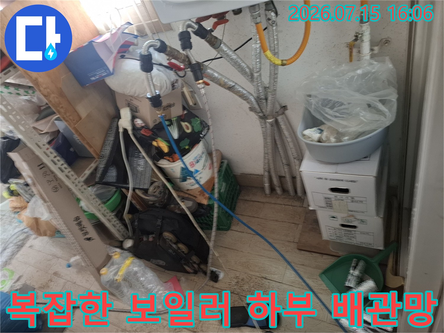 은색 주름관과 파란색 호스가 연결된 보일러 하부-주변에 짐들이 놓여 있는 현장 사진