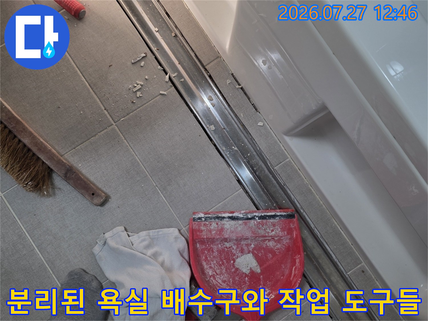 회색 타일 바닥 위에 탈거된 은색 트랜치 배수구가 놓여 있고 청소용품들이 주변에 배치된 사진