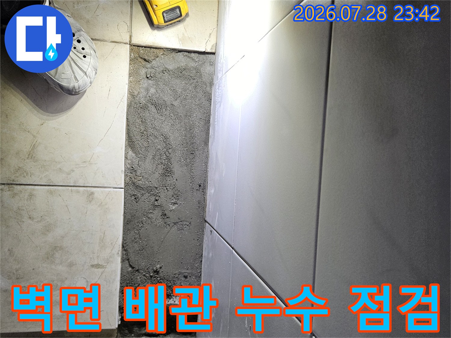 전동공구와 크록스를 착용한 작업자가 벽면의 노출된 배관 부위를 점검하는 모습