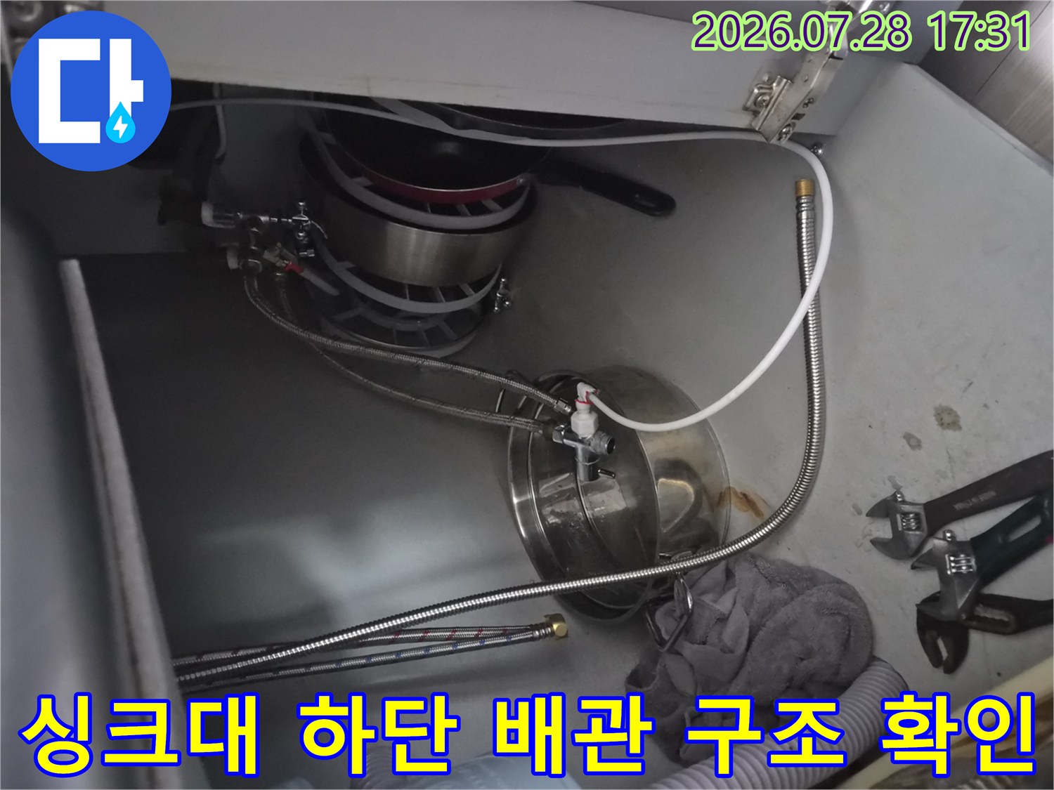 스테인리스 소재의 배관과 금속 호스가 얽힌 싱크대 아래쪽 모습입니다.