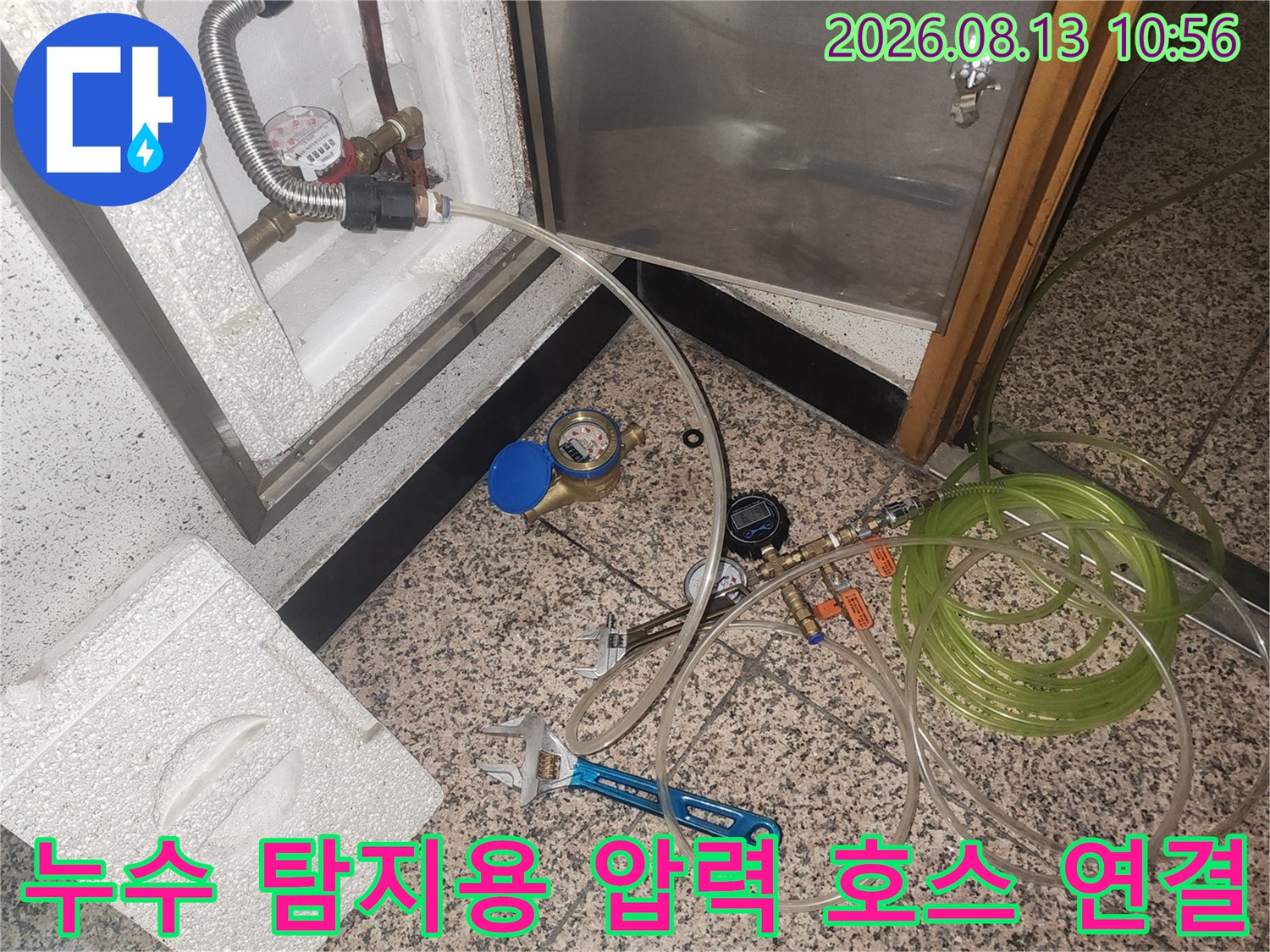 흰색 점검구 내부의 금속 배관에 투명한 호스와 측정기가 연결되어 있는 모습-작업 도구가 바닥에 배치됨